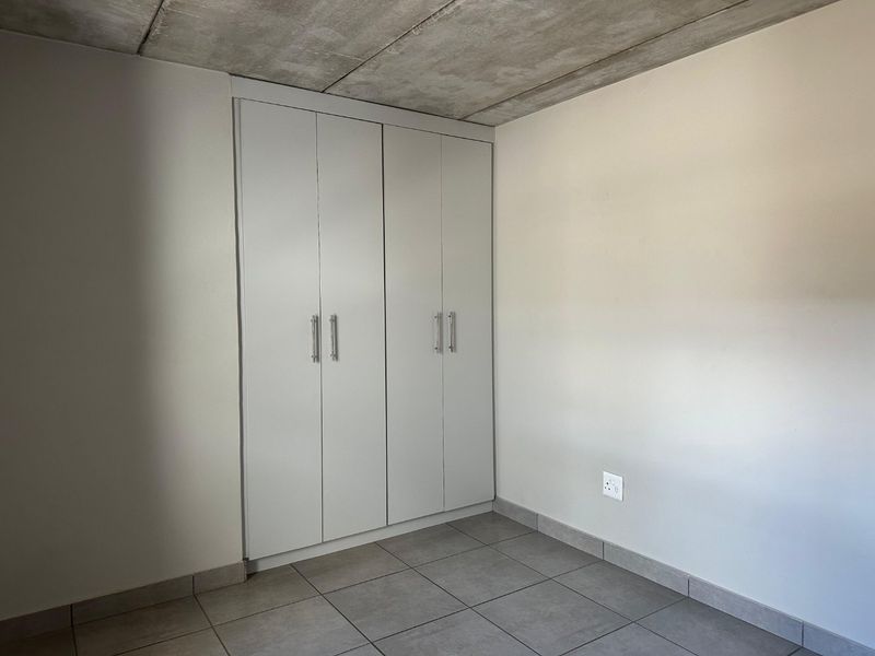 A clean, modern bedroom featuring built-in cupboards, neutral tiled flooring, and an industrial-style exposed concrete ceiling.