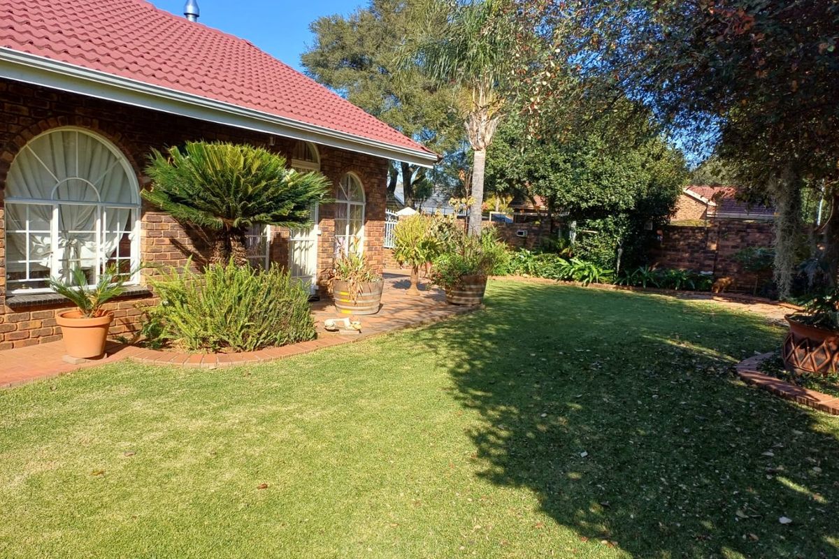 A well-maintained rear garden featuring a brick facade, arched windows, and a paved patio area, surrounded by established landscaping and a brick boundary wall.