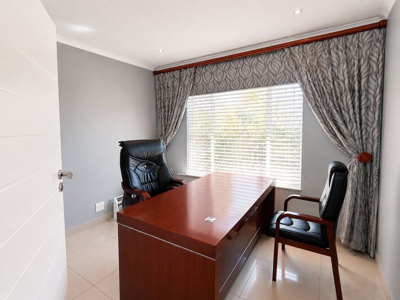A well-maintained home office featuring neutral wall tones, tiled flooring, recessed lighting, and large windows with fitted blinds and curtains.