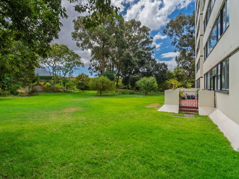A view of the apartment building's rear facade featuring a well-maintained lawn and landscaped garden area with direct access via a security gate.