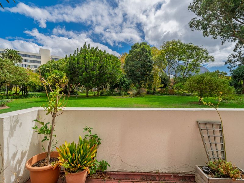 A private balcony featuring a concrete balustrade and tiled flooring, overlooking a lush, well-maintained communal garden area.