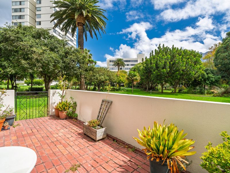 A private ground-floor patio featuring red brick paving and a low boundary wall, overlooking a shared garden area with mature trees.