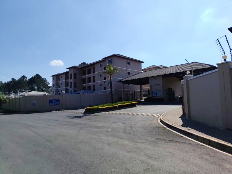 The entrance to the Royal Reef apartment complex, featuring a secure gatehouse, perimeter wall with electric fencing, and well-maintained paved access roads.