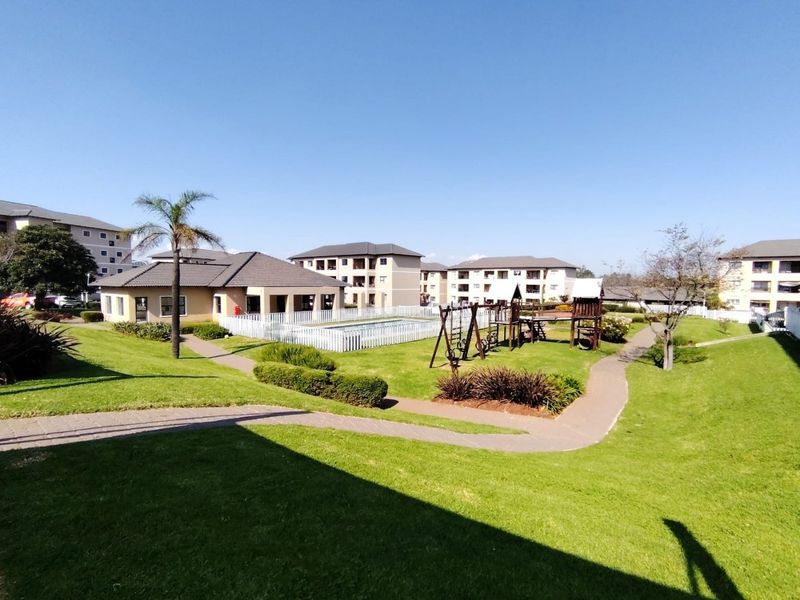 Communal outdoor area featuring a swimming pool with safety fencing, landscaped lawns, paved pathways, and a playground set within an apartment complex.