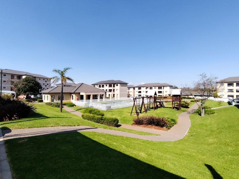 A well-maintained communal garden area featuring paved walkways, a swimming pool, and playground facilities within an apartment complex.