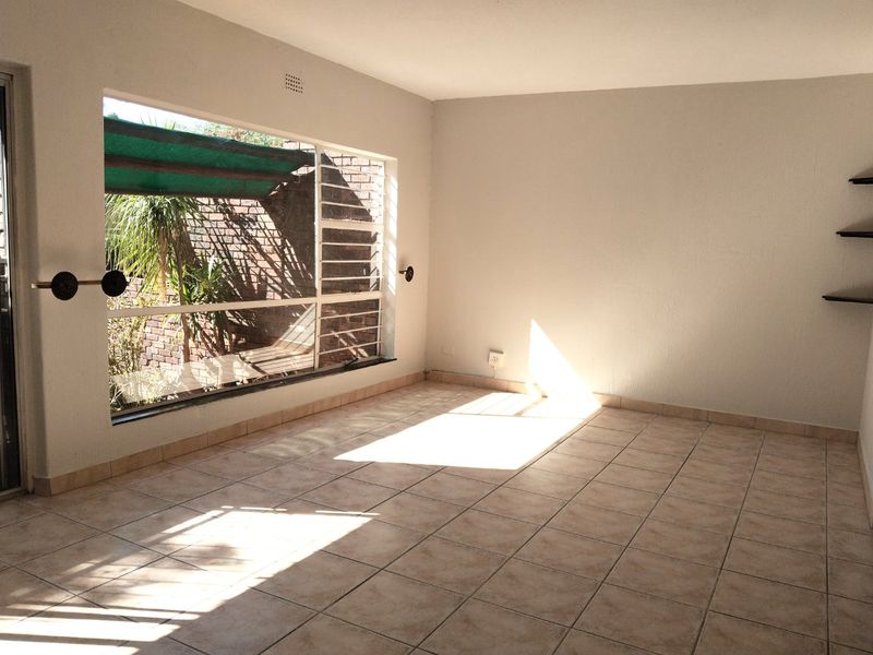 A bright, empty living room featuring tiled flooring, large windows with security bars, and built-in floating wall shelves.