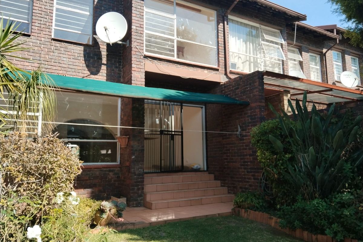 Rear view of a face brick townhouse featuring a small garden, tiled entrance steps, security gate, and an awning over the patio area.