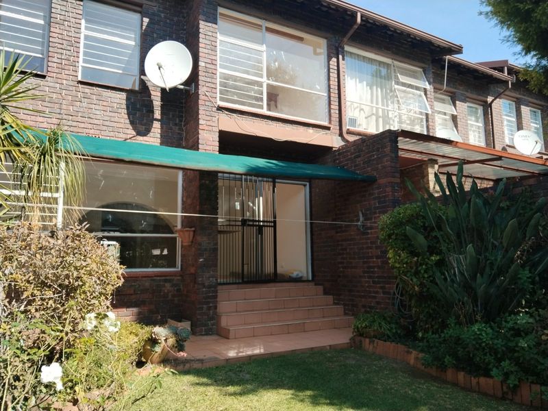 Rear view of a face brick townhouse featuring a small garden, tiled entrance steps, security gate, and an awning over the patio area.