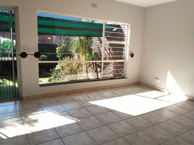 A bright, empty living area featuring tiled flooring, a large window with security bars, and direct access to an outdoor patio space.