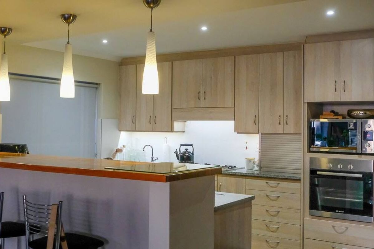 Modern kitchen featuring light wood cabinetry, stone countertops, a timber breakfast bar, and integrated appliances including an oven and microwave.