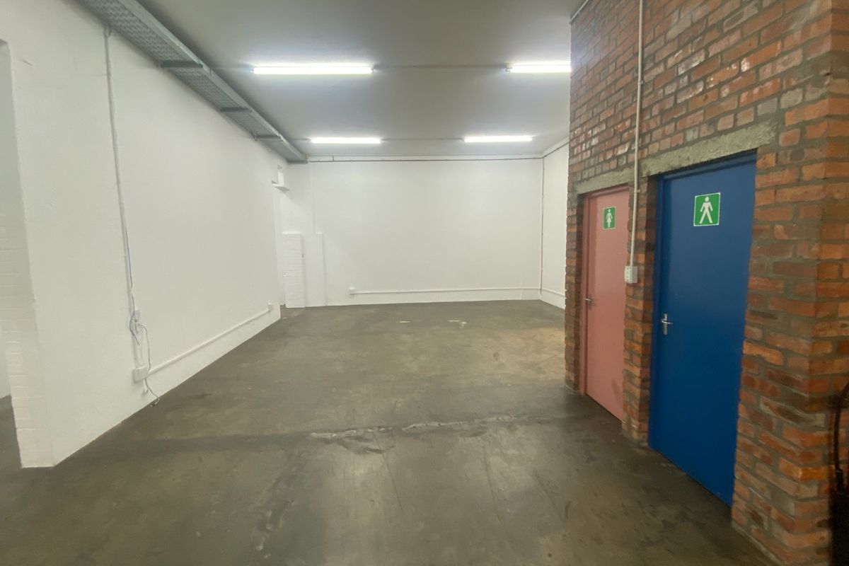Open-plan industrial utility space featuring exposed brickwork, concrete flooring, fluorescent lighting, and dual access doors leading to restroom facilities.