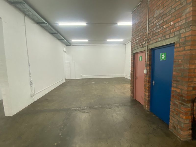 Open-plan industrial utility space featuring exposed brickwork, concrete flooring, fluorescent lighting, and dual access doors leading to restroom facilities.
