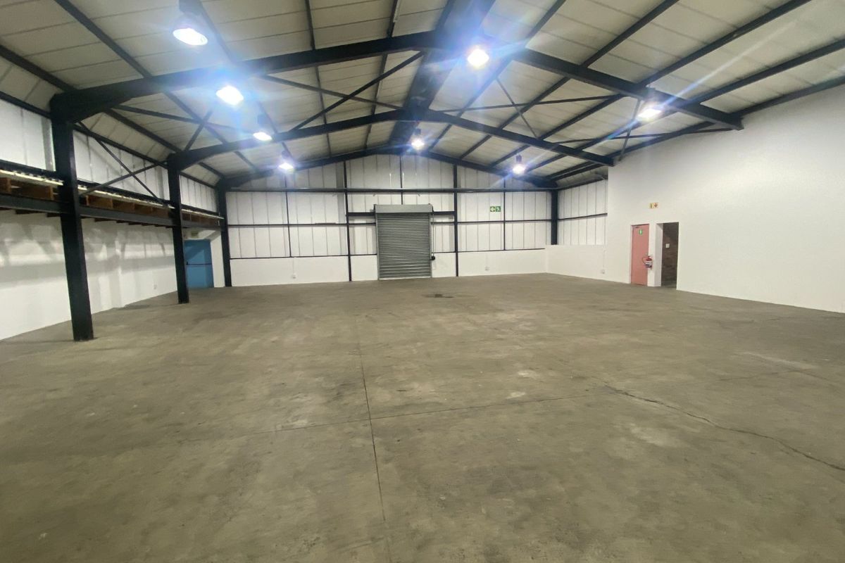 Spacious industrial warehouse unit featuring high ceilings, exposed steel trusses, concrete flooring, and a large roller shutter door for easy access.