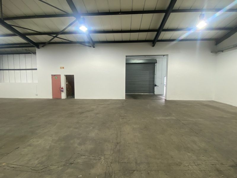 Spacious industrial warehouse unit featuring high ceilings, exposed steel trusses, concrete flooring, and a large roller shutter door for vehicle access.