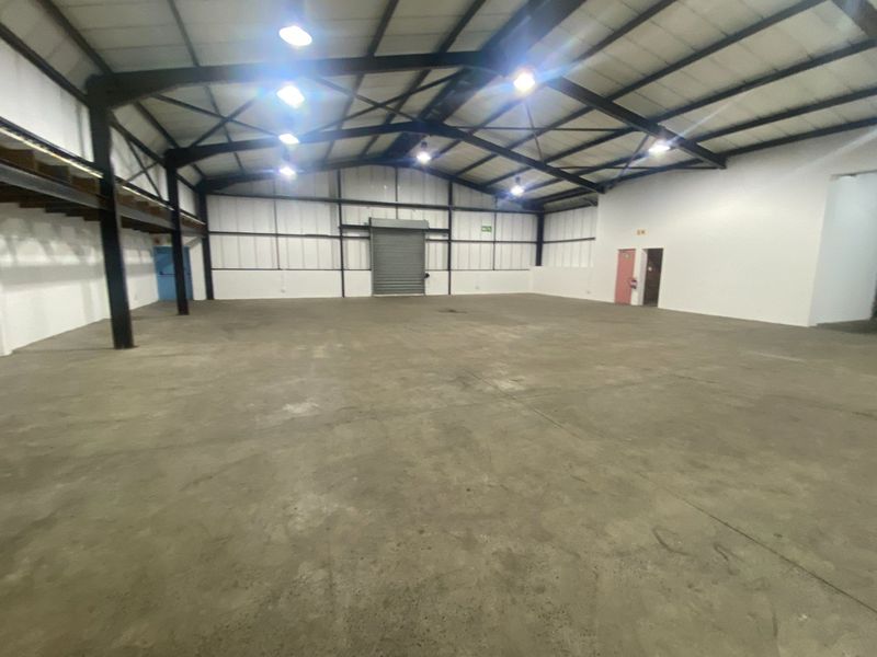 Spacious industrial warehouse featuring high ceilings, exposed steel trusses, concrete flooring, and a large roller shutter door for easy access.