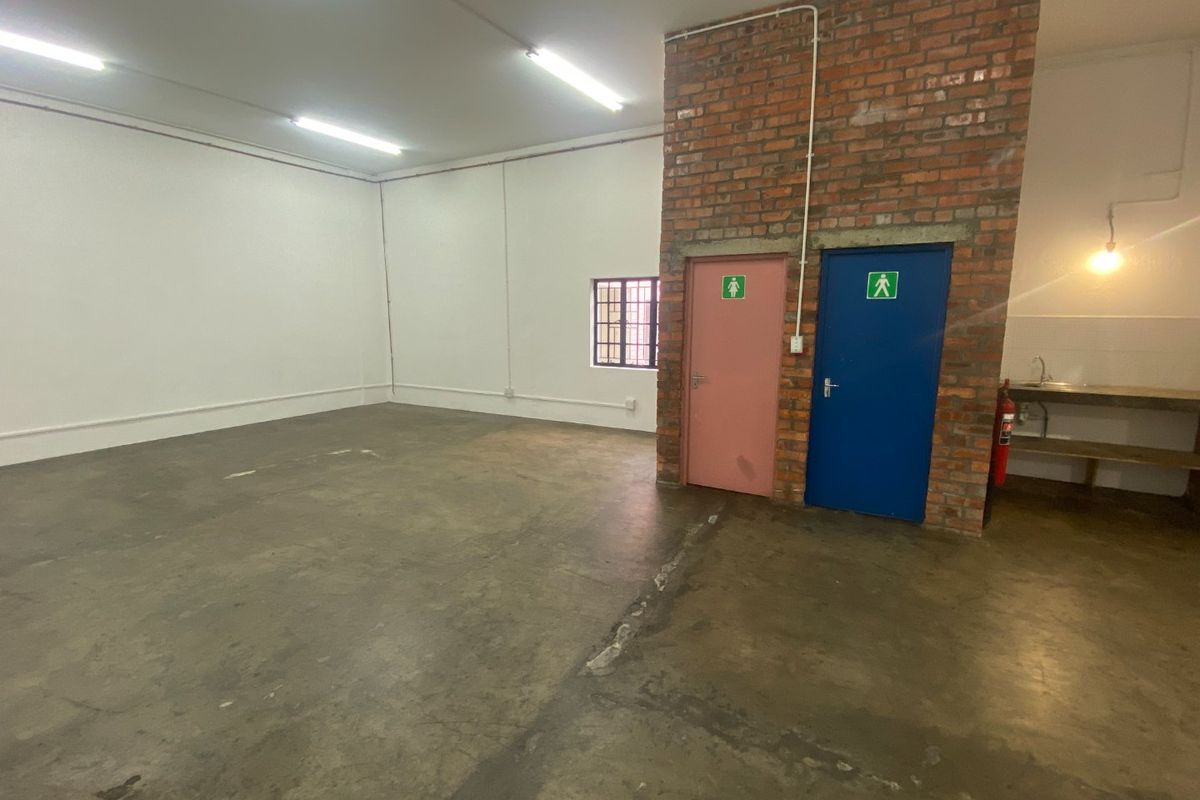 Open-plan industrial warehouse space featuring exposed brickwork, concrete flooring, fluorescent lighting, and dedicated restroom access.