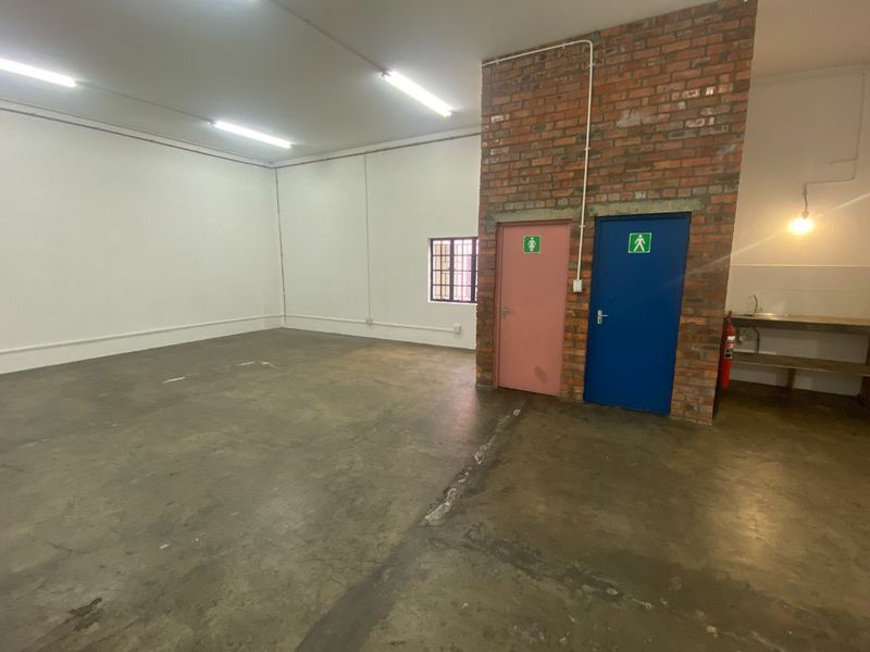 Open-plan industrial warehouse space featuring exposed brickwork, concrete flooring, fluorescent lighting, and dedicated restroom access.