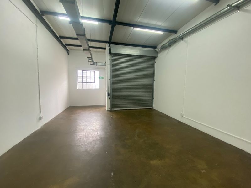 A clean, empty industrial utility space featuring a roller shutter door, exposed ducting, and durable concrete flooring, suitable for storage or workshop use.
