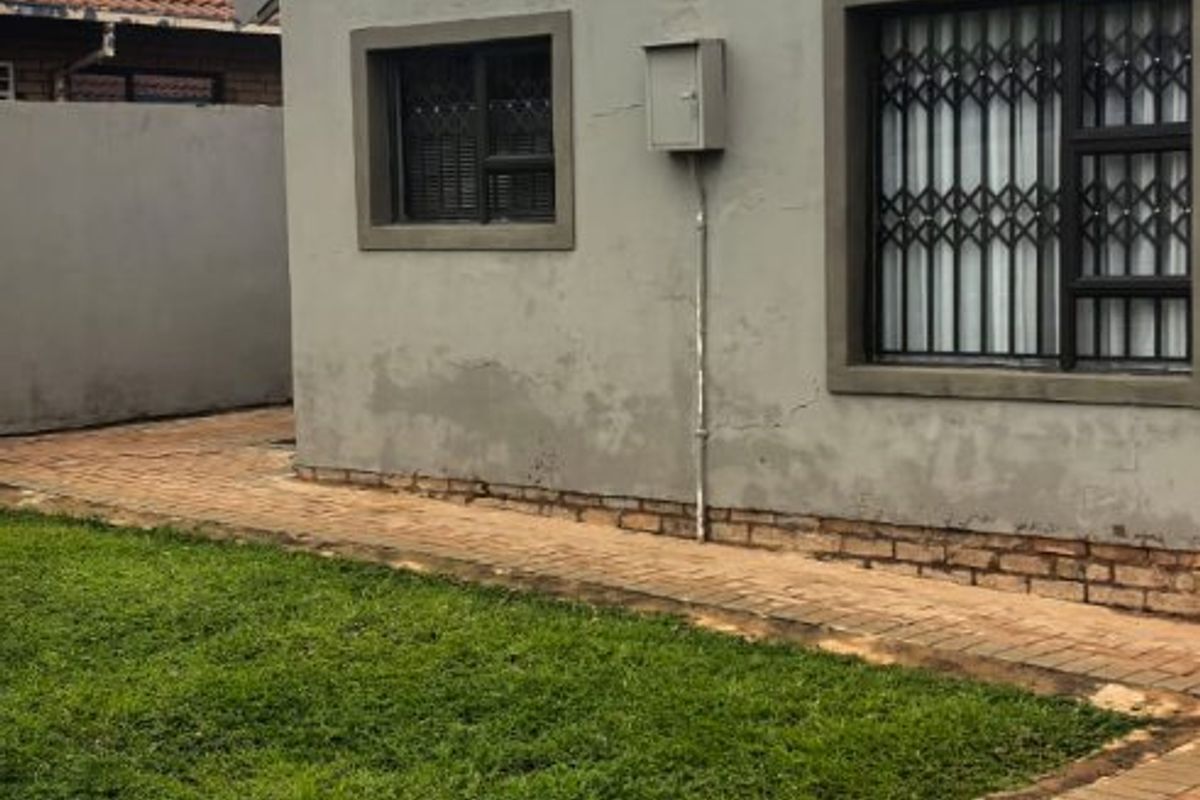 Rear exterior view of the house featuring a paved walkway, small lawn, security gates on windows, and visible exterior wall maintenance requirements.