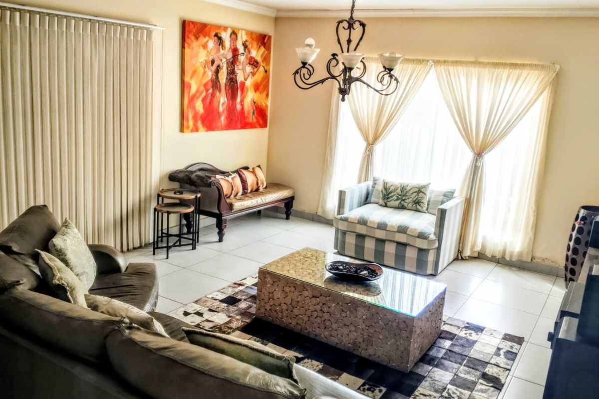 A spacious living room featuring neutral tiled flooring, a decorative wrought iron chandelier, and large windows dressed with vertical blinds and sheer curtains.