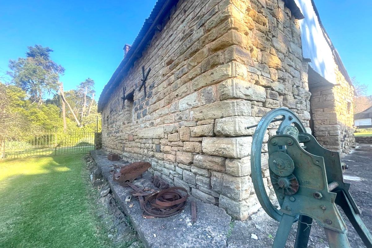 Exterior view of a rustic stone farm building featuring traditional masonry, a corrugated metal roof, and a concrete foundation plinth set within a garden.
