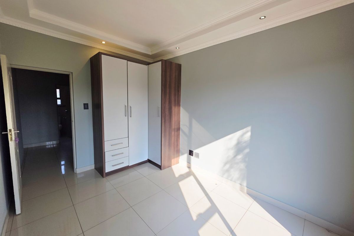 A bright, empty bedroom featuring neutral tiled flooring, recessed ceiling lighting, decorative coved cornicing, and a modern built-in wardrobe with a two-tone finish.