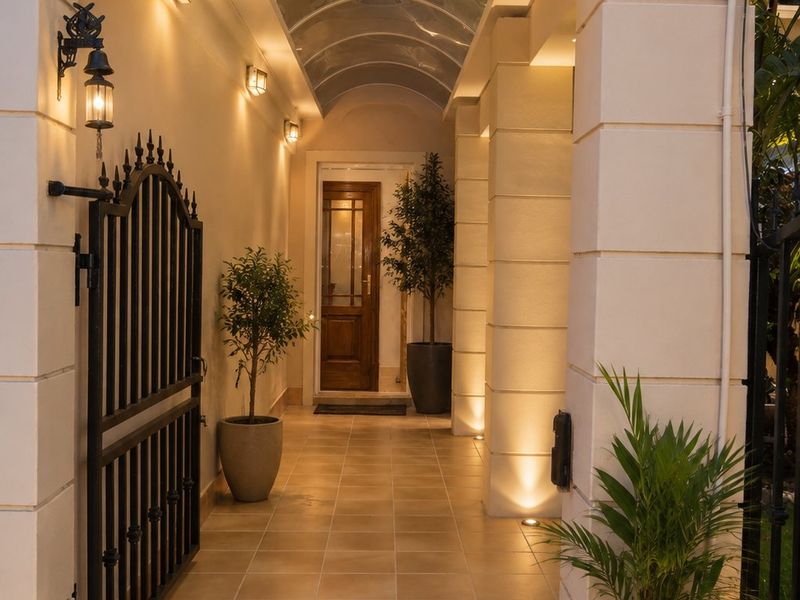 A well-lit, covered entrance walkway featuring tiled flooring, an arched polycarbonate roof, and a secure wrought iron gate leading to a timber front door.