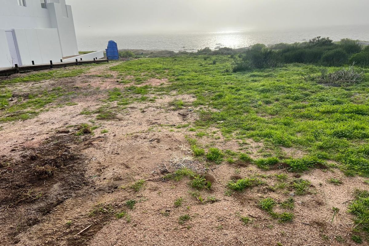 A vacant residential plot featuring natural coastal vegetation and direct ocean views, situated adjacent to an existing white building structure.