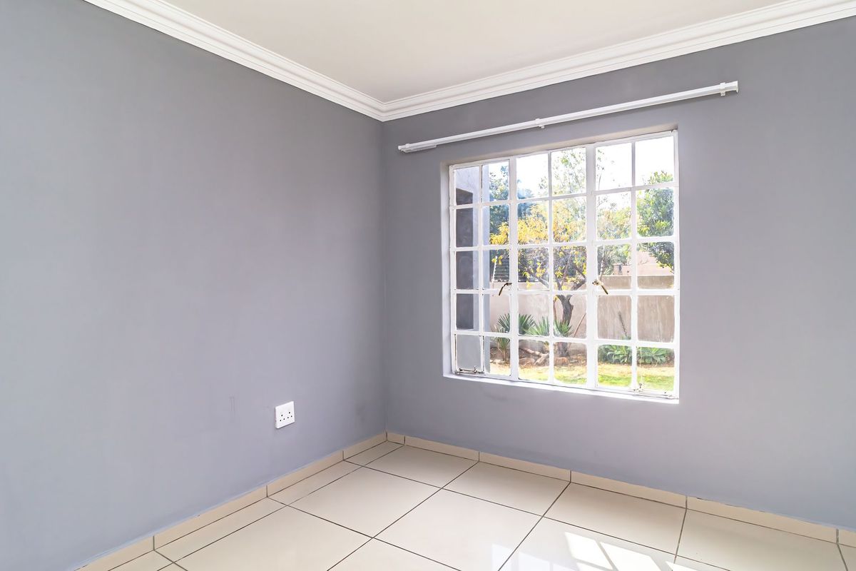 A bright, empty bedroom featuring neutral grey walls, tiled flooring, crown molding, and a large lattice window overlooking the garden.