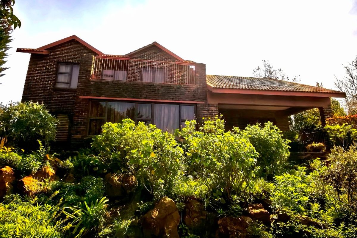 A rear view of a double-story brick house featuring a balcony with metal railings, a covered patio area, and established garden landscaping.