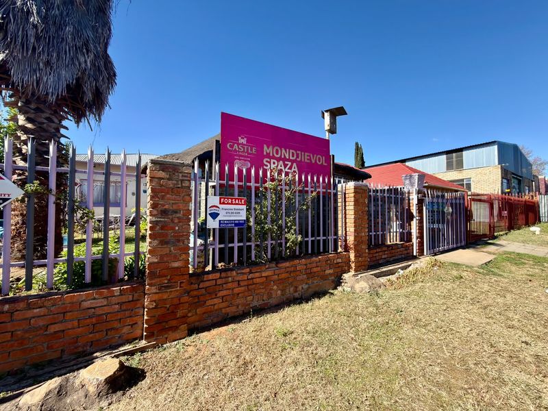 Front exterior view of the property featuring a brick boundary wall, metal security fencing, and a combination of thatched and metal roofing.