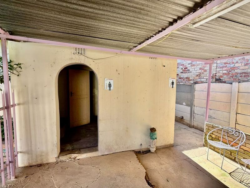 A rear exterior area featuring a covered concrete patio, arched doorway, and brick boundary wall, showing signs of significant wear and weathering.