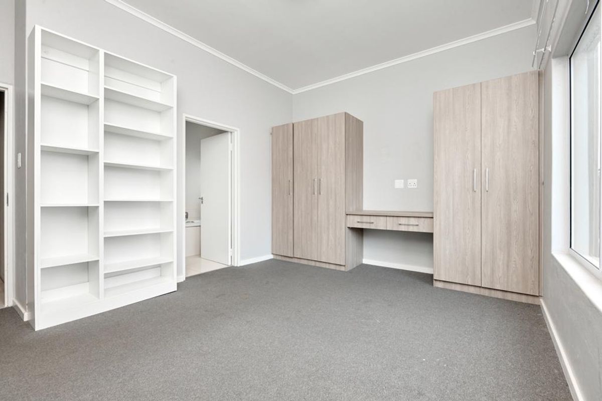 A bright, empty bedroom featuring neutral grey carpeting, built-in wooden cabinetry with an integrated desk, and a large white shelving unit.