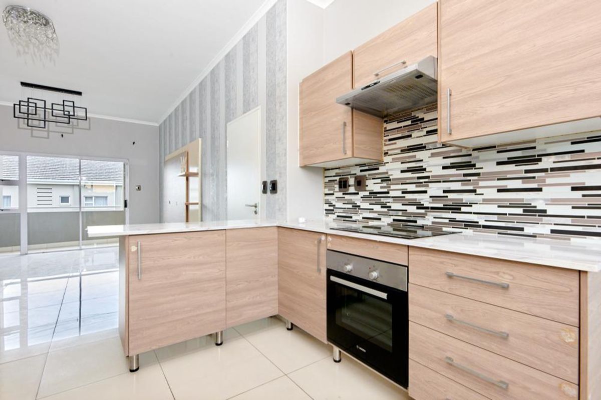 Modern kitchen featuring light wood-finish cabinetry, stone countertops, mosaic tile backsplash, and integrated appliances, flowing into an open-plan living area with balcony access.