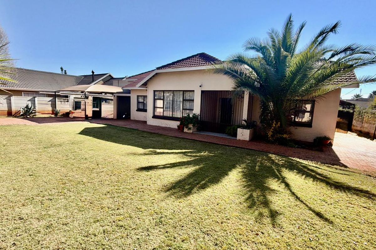 A well-maintained rear exterior view featuring a spacious lawn, paved driveway, and a covered patio area with a shade sail.