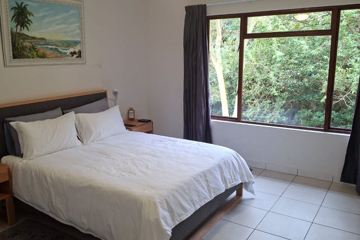A bright bedroom featuring tiled flooring, a ceiling fan, and a large window offering a pleasant view of the surrounding greenery.