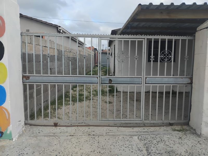 Front view of the property featuring a metal security gate, concrete boundary wall, and a basic carport structure over the driveway area.