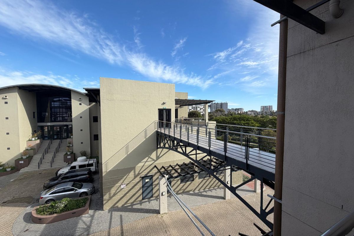 Exterior view of a modern commercial building complex featuring a raised metal pedestrian walkway connecting two building sections over a paved courtyard.