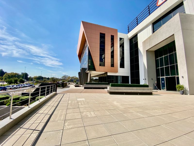 Modern commercial building exterior featuring a spacious paved terrace, contemporary metal and glass facade, and integrated concrete seating areas.