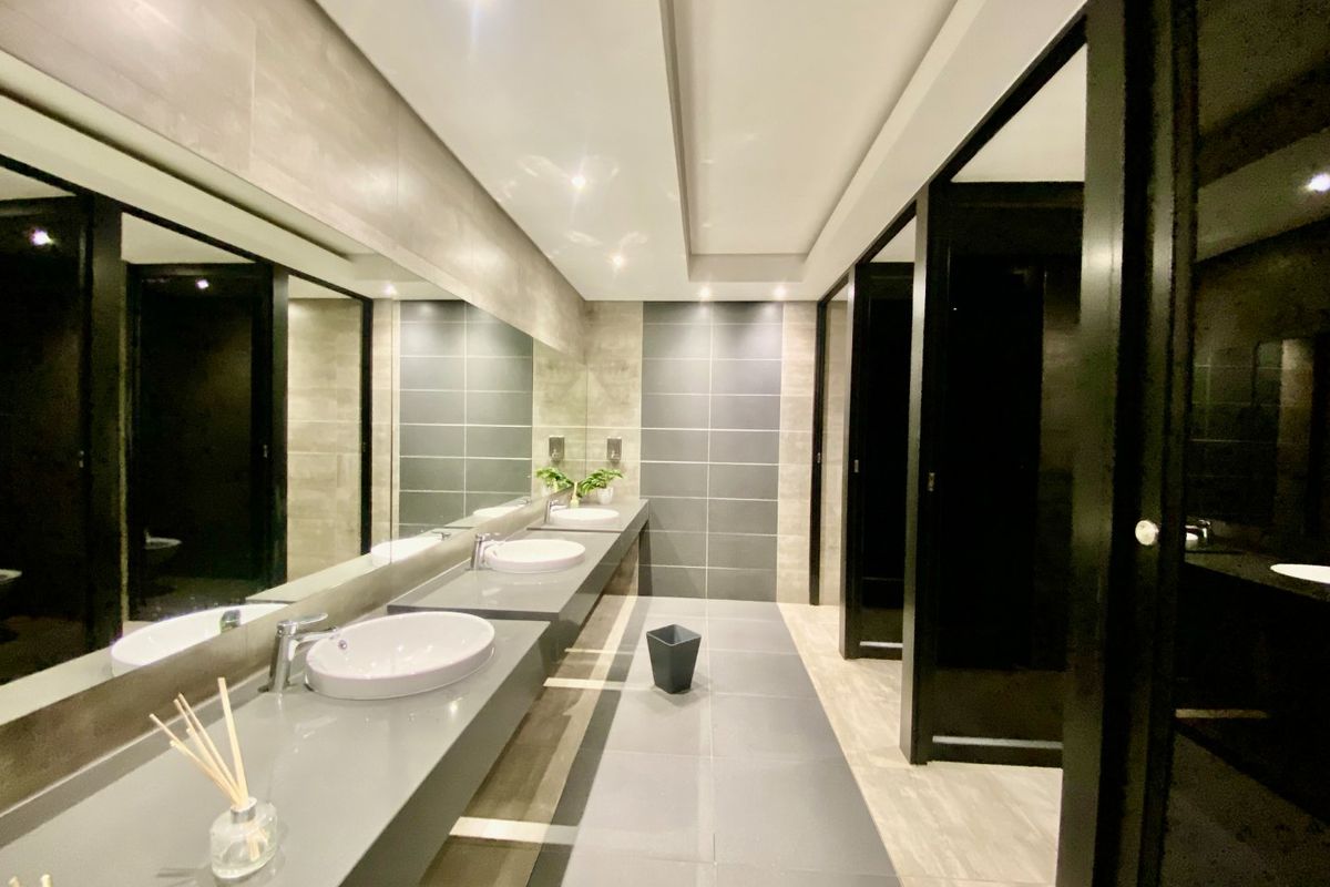 Modern commercial bathroom featuring a sleek floating vanity with dual vessel sinks, large wall-to-wall mirror, recessed lighting, and private partitioned toilet cubicles.