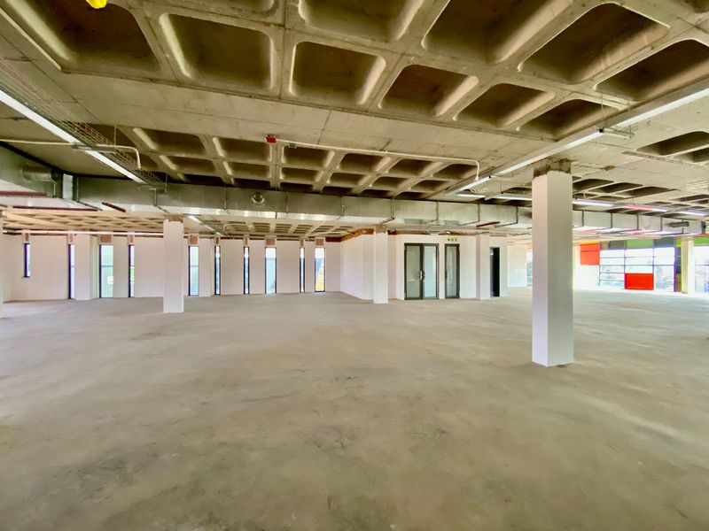 A large, open-plan commercial office space featuring an exposed waffle slab ceiling, industrial HVAC ducting, and expansive windows providing ample natural light.