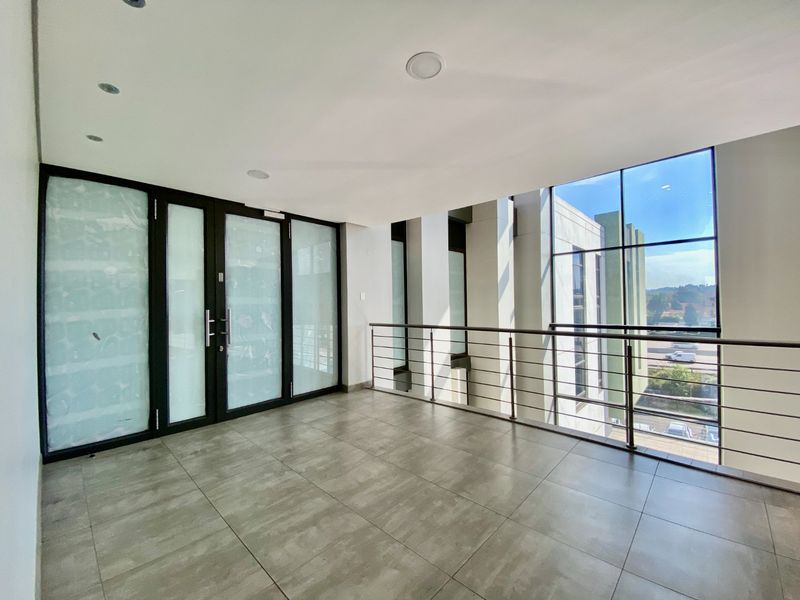 A bright, modern commercial hallway featuring large glass windows, tiled flooring, and a stainless steel balustrade overlooking the exterior.