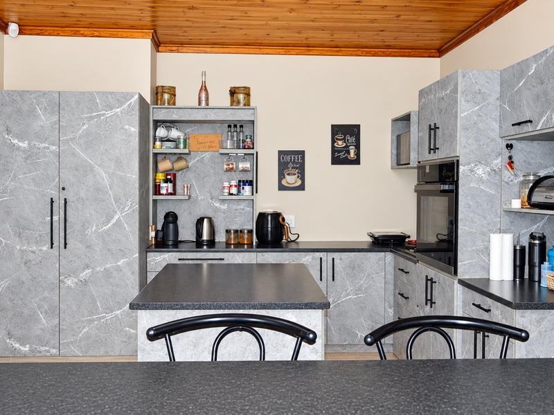 A modern kitchen featuring grey marble-effect cabinetry, dark laminate countertops, a central island, and a warm timber-paneled ceiling.