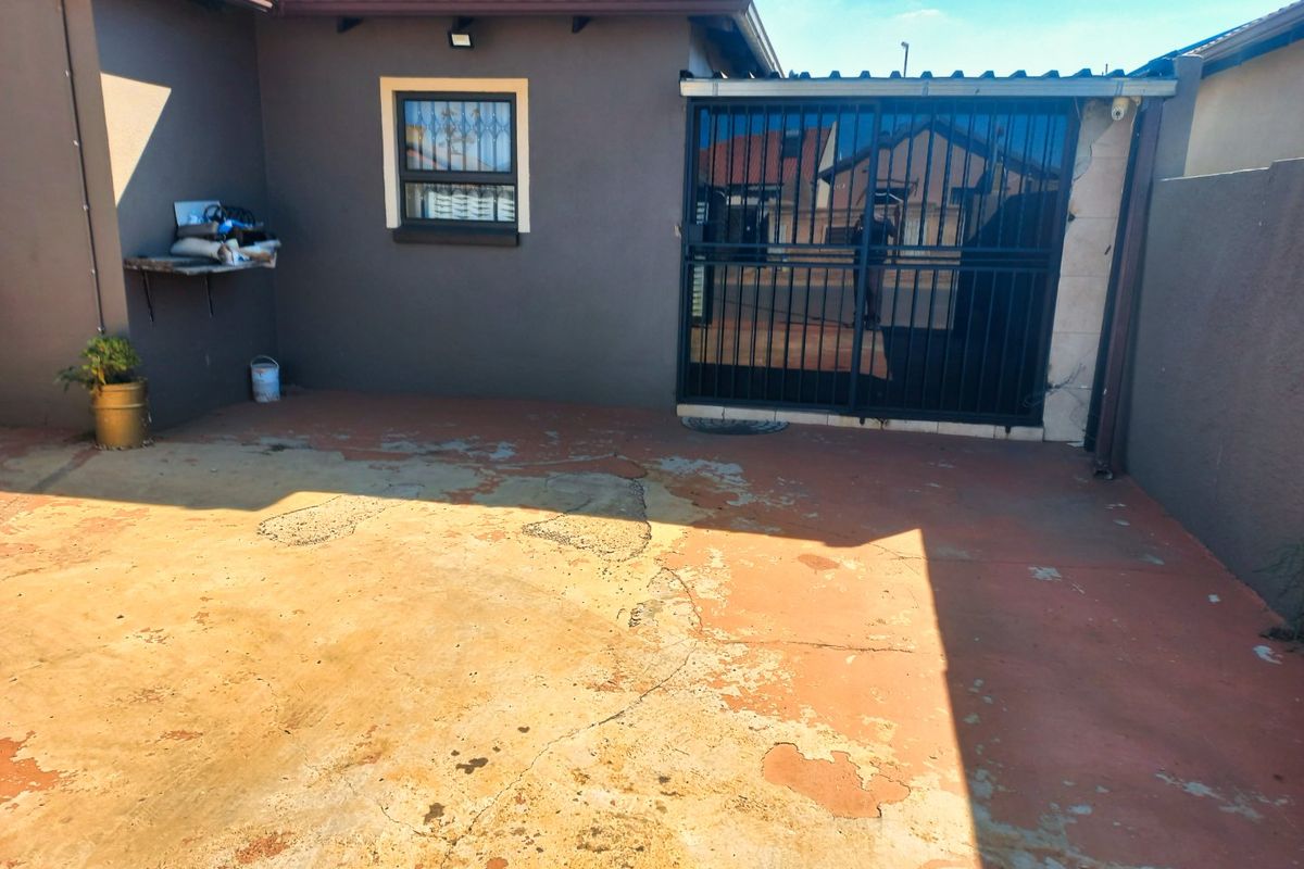A rear exterior area featuring a security gate and concrete paving, showing signs of wear and requiring maintenance to the surface and wall finishes.