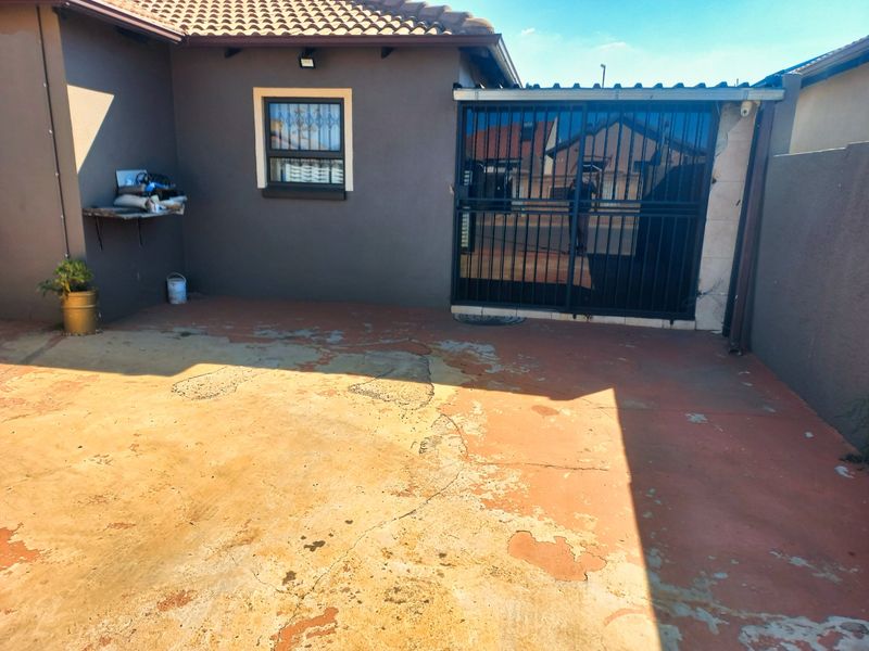 A rear exterior area featuring a security gate and concrete paving, showing signs of wear and requiring maintenance to the surface and wall finishes.