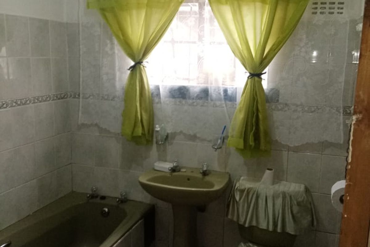 A functional bathroom featuring a bathtub, pedestal basin, and toilet with neutral wall tiling and security bars on the window.