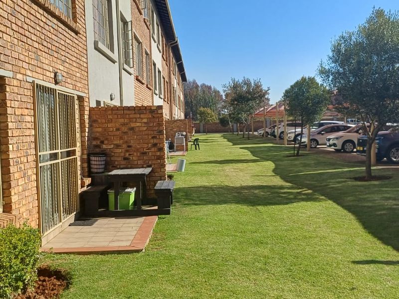 Rear view of the apartment complex featuring face brick exterior, private paved patio areas, and well-maintained communal lawn with adjacent carport parking.