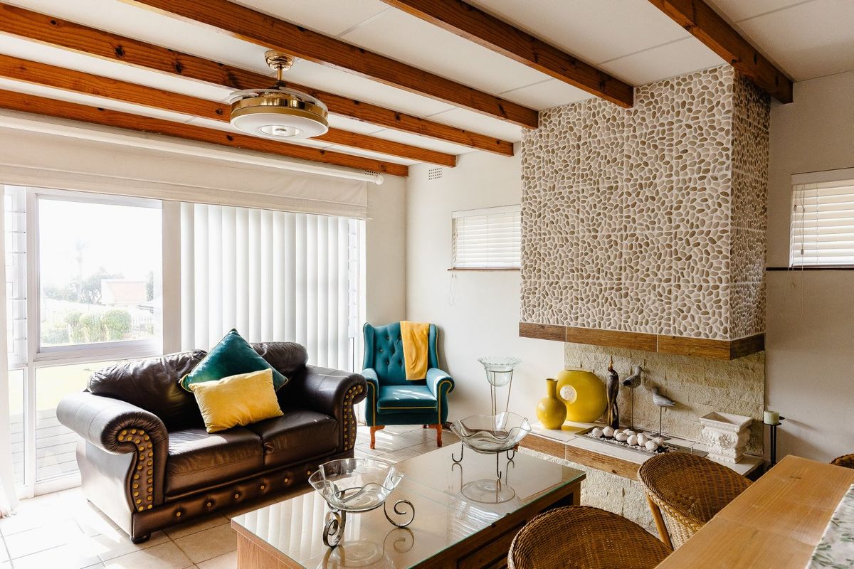 A living room featuring exposed timber ceiling beams, a distinctive pebble-clad fireplace, tiled flooring, and large windows providing natural light.