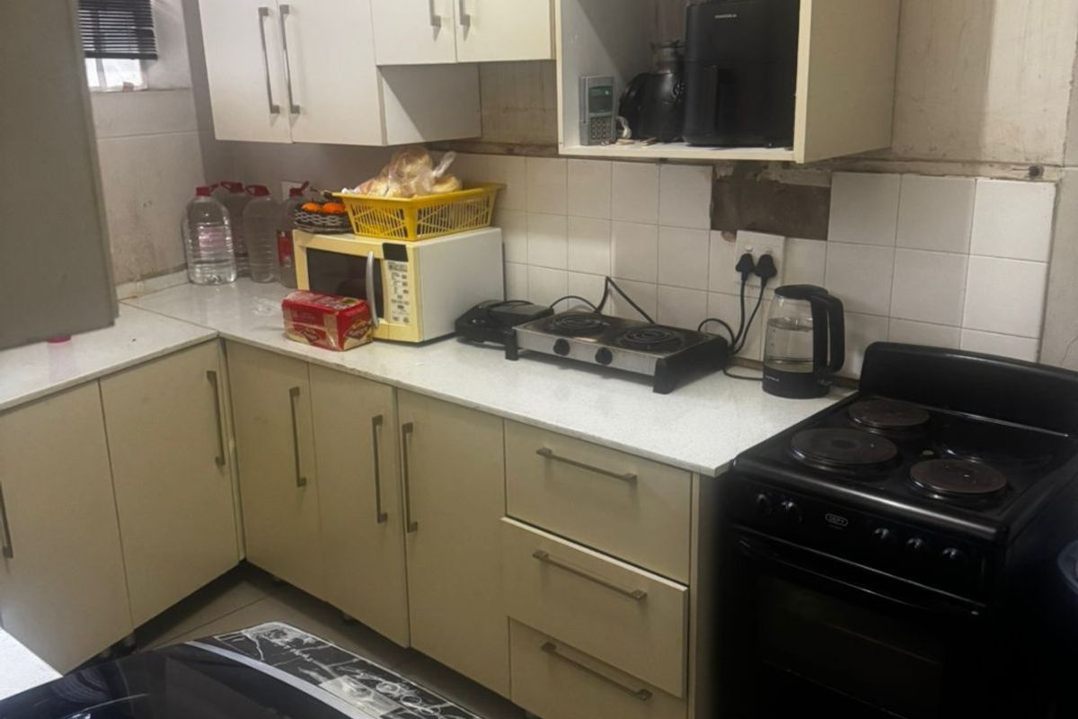 A compact kitchen featuring laminate cabinetry, stone-look countertops, and a tiled splashback, currently showing signs of wear and requiring maintenance to the wall finishes.