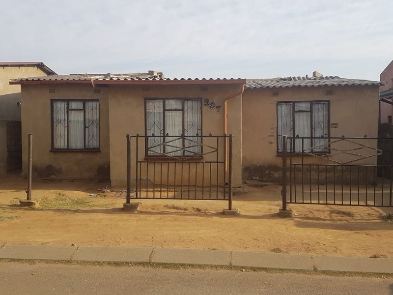 Front elevation of a single-story house featuring a corrugated roof, metal security window bars, and a fenced yard area.